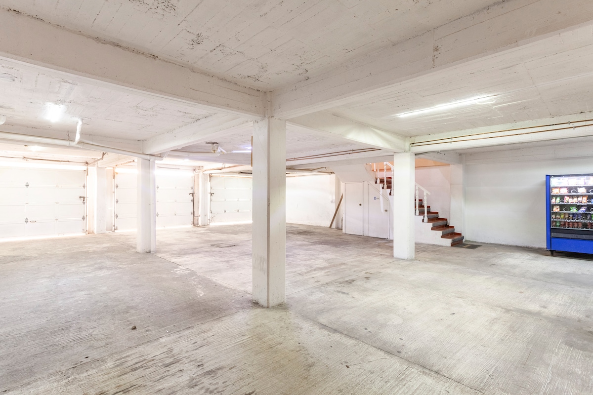 A spacious parking area features multiple garage doors for vehicle access, accompanied by white walls and a concrete floor. A staircase is visible in the background, offering access to upper levels. A vending machine can be seen along one wall, providing snacks and beverages.
