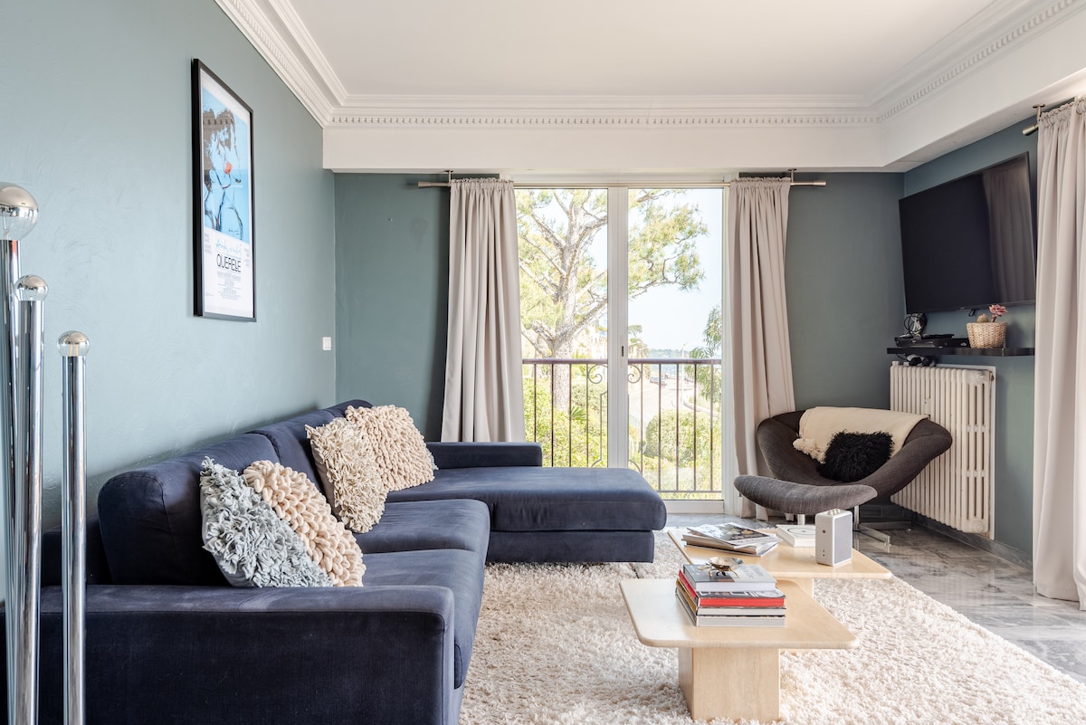 A spacious living area is highlighted by a dark sofa and a contemporary armchair. Natural light flows through the floor-to-ceiling sliding doors, which open to a lovely view. The room features neutral tones, a light area rug, a wooden coffee table, and a mounted television.