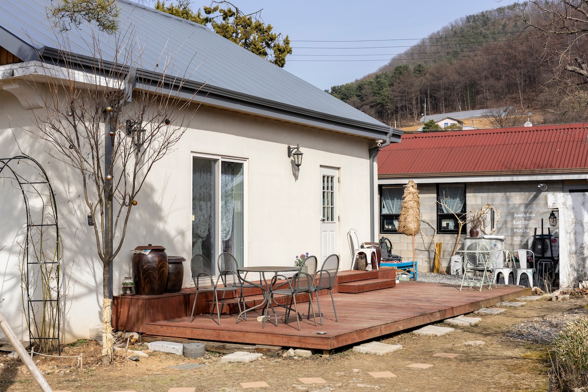 A cozy outdoor deck features a wooden surface with a round table and several chairs, surrounded by a tranquil garden area. The serene facade of a white house is visible, complemented by decorative elements and natural landscaping. A neighboring structure adds to the inviting atmosphere.