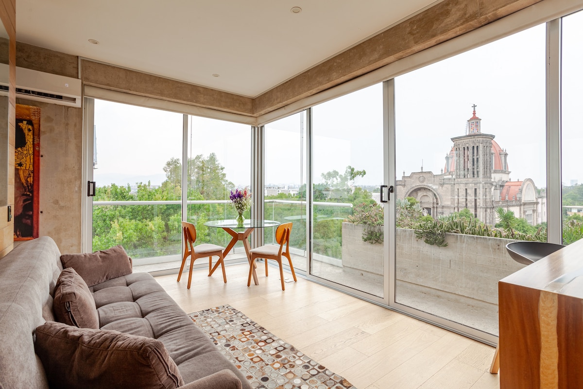 A modern living space features large glass doors that offer a panoramic view of greenery and an architectural landmark. A comfortable gray sofa is positioned next to a small dining area with two chairs and a wooden table. Natural light fills the room.