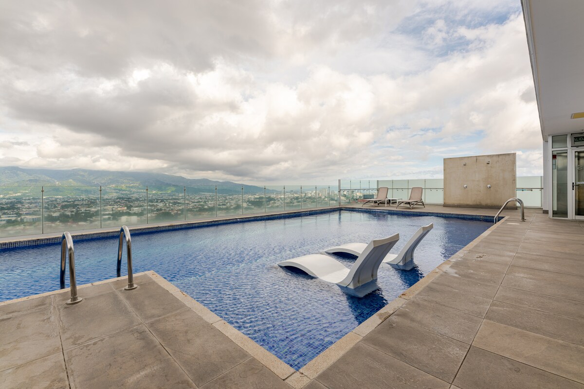 A rooftop pool is visible, featuring sleek lounge chairs partially submerged in the inviting water. The spacious area showcases modern tiling and stunning views of the surrounding mountains and cityscape under a partially cloudy sky.