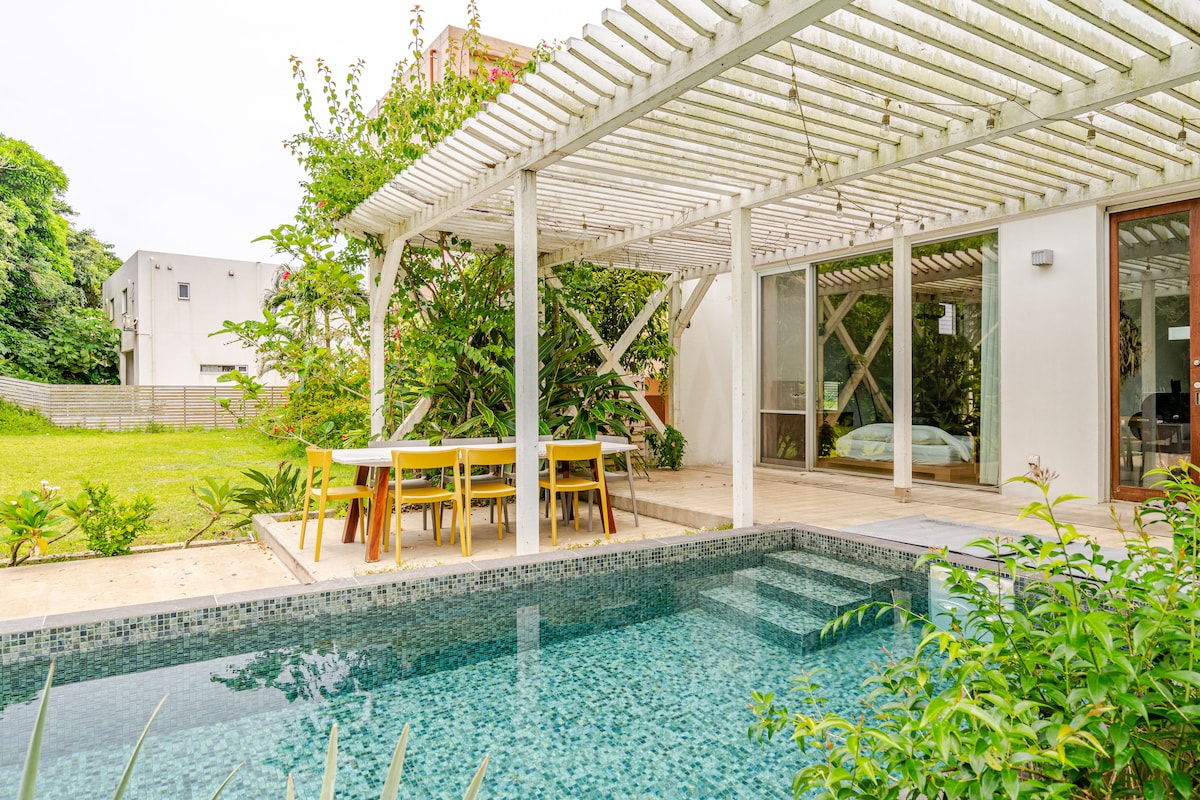 An outdoor area is highlighted by a refreshing pool surrounded by a tiled edge. A wooden pergola provides shade over a dining table with yellow chairs. Lush greenery and a spacious garden are visible in the background, enhancing the serene setting.