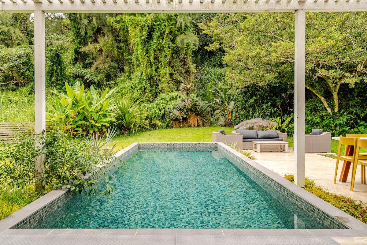A cooling swimming pool is framed by lush greenery and outdoor seating. The tranquil water reflects the surrounding tropical plants, while a shaded terrace provides a comfortable spot for relaxation. Stone tiles lead to a patio area featuring a modern dining set and cozy lounge furniture.