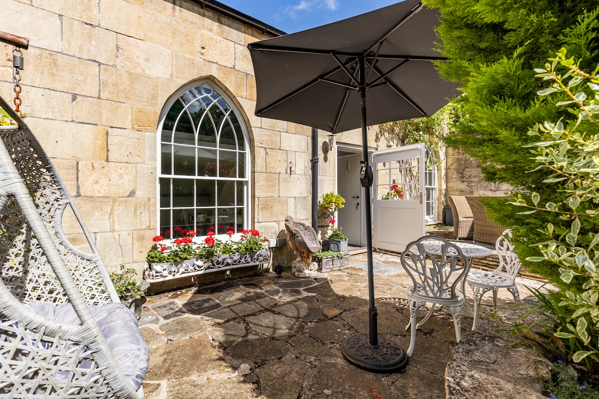 An outdoor seating area is highlighted by a round table and two chairs, positioned beneath a large umbrella. Vibrant flowers are arranged in window boxes along a stone wall, while a comfortable hanging chair invites relaxation. Lush greenery frames the space, creating a peaceful environment.