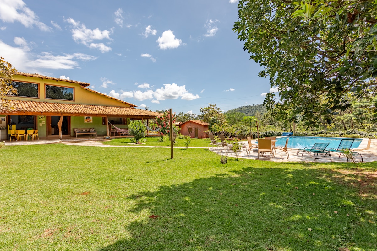 A vibrant green yard features a spacious house with a tile roof, surrounded by trees. A swimming pool is nestled beside the patio, where lounge chairs are arranged. The outdoor space offers a relaxing atmosphere, ideal for family gatherings or quiet retreats.