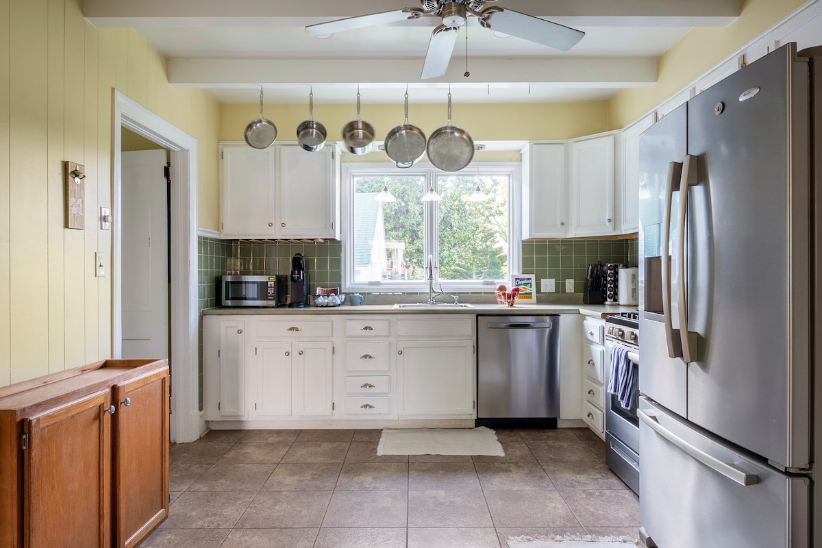 A spacious kitchen is shown, featuring white cabinetry and a stainless steel refrigerator. A large window provides natural light, illuminating the countertops. Cooking essentials include a microwave and a dishwasher. A ceiling fan offers airflow, while hanging pots add a functional touch.
