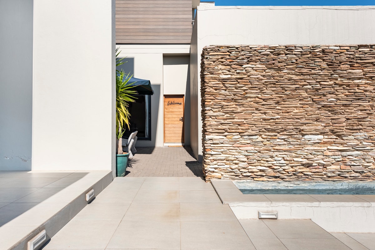 A modern entrance showcases a stylish stone wall alongside a serene water feature. Invisible in the image are welcoming outdoor seating areas. A clean pathway leads to a wooden door, indicating access to the cottage, set against a bright blue sky.