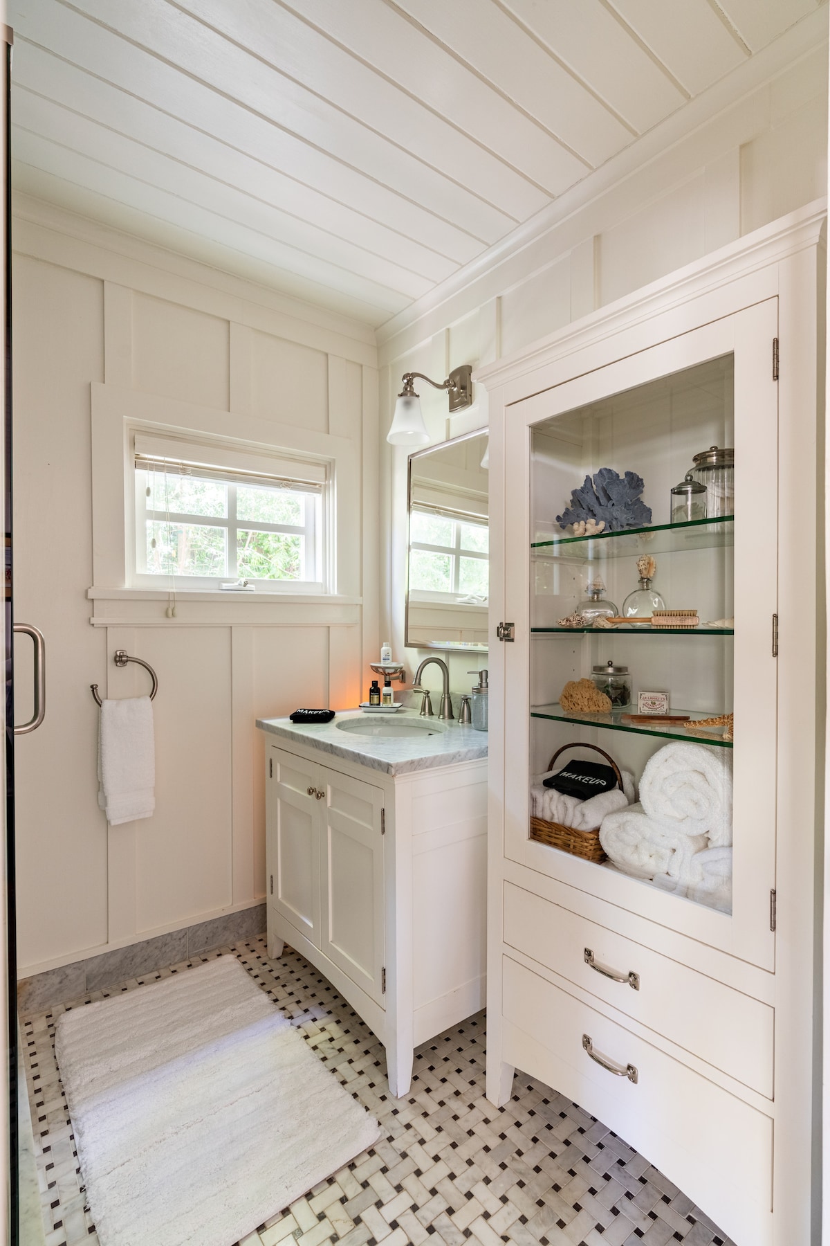 A clean and well-appointed bathroom features light-colored wooden paneling and marble flooring. A modern vanity with a sink is positioned beneath a large mirror, while a glass-front cabinet displays neatly arranged towels and decorative items. Natural light filters through a small window.