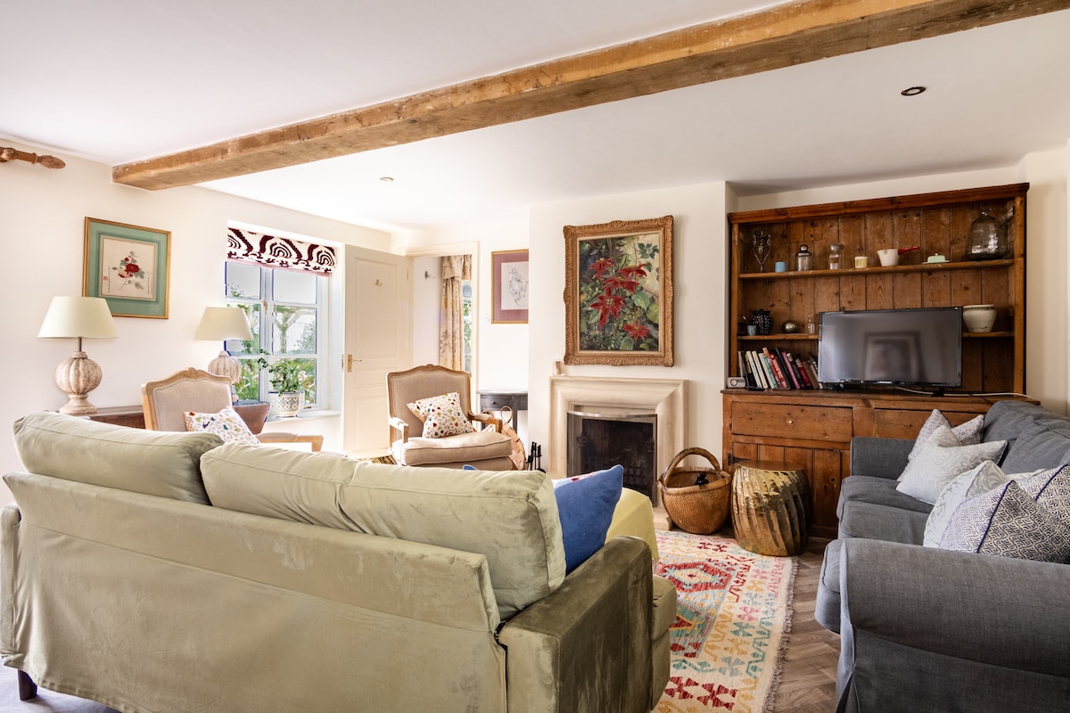 A spacious sitting room is furnished with cozy sofas and armchairs arranged around a coffee table. A wooden beam extends across the ceiling, while a fireplace and a TV unit provide focal points. Natural light filters in through the windows, creating a welcoming space.