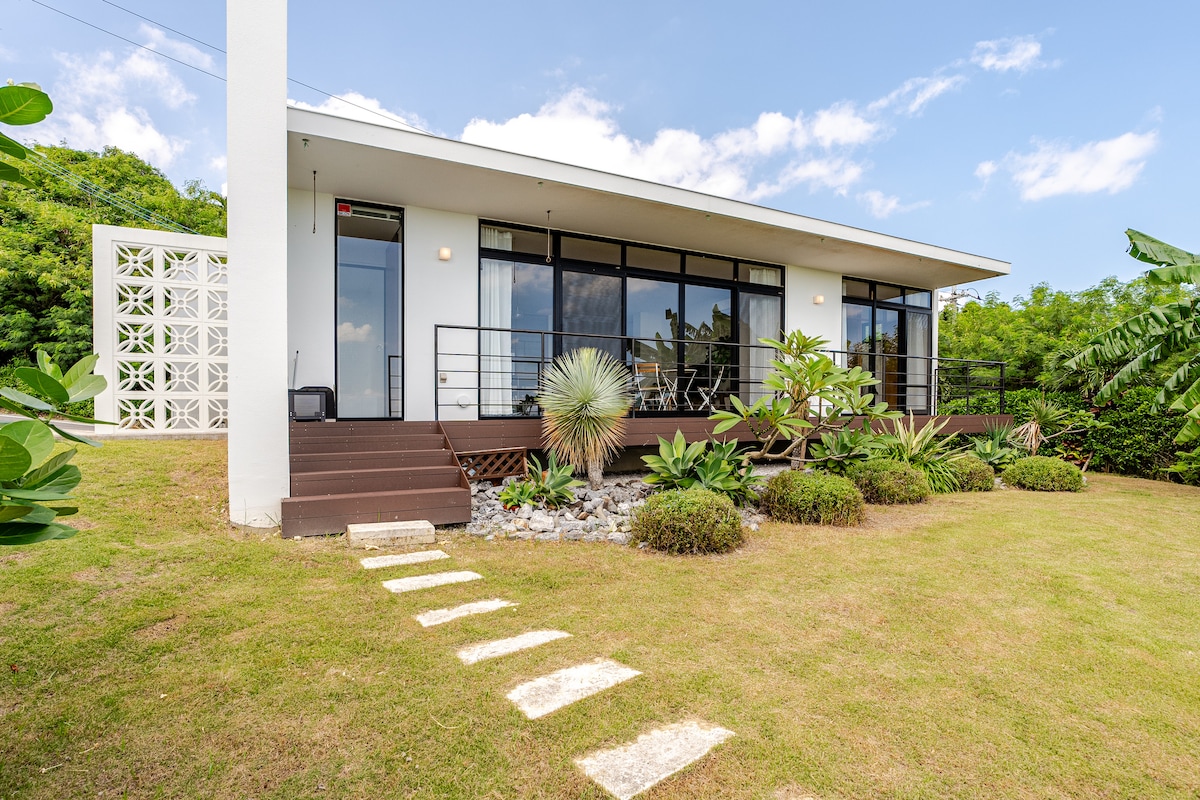 A modern single-story house features large glass windows that overlook a landscaped garden. Tropical plants and greenery surround the exterior, creating a serene environment. A stone pathway leads to the entrance, emphasizing the natural beauty of the property.