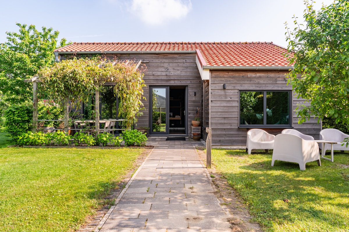 The cottage's exterior is highlighted by a wooden façade and a red tiled roof, surrounded by lush greenery. A paved pathway leads to the entrance, with a seating area featuring white chairs, and a pergola adorned with climbing plants providing shade.