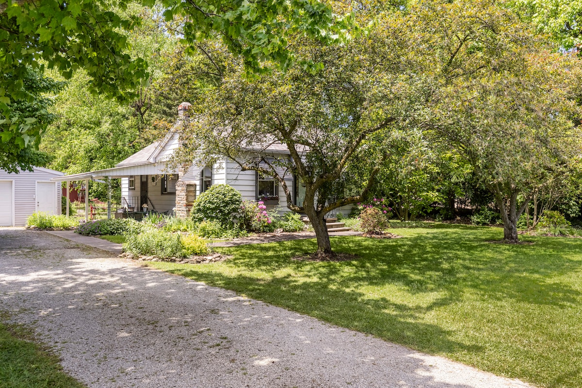 A one-story mid-century cottage is set among lush greenery. The property features a covered porch, surrounded by vibrant flower beds and large trees, offering a shaded area. A gravel driveway leads to the cottage, contributing to the rural ambiance.