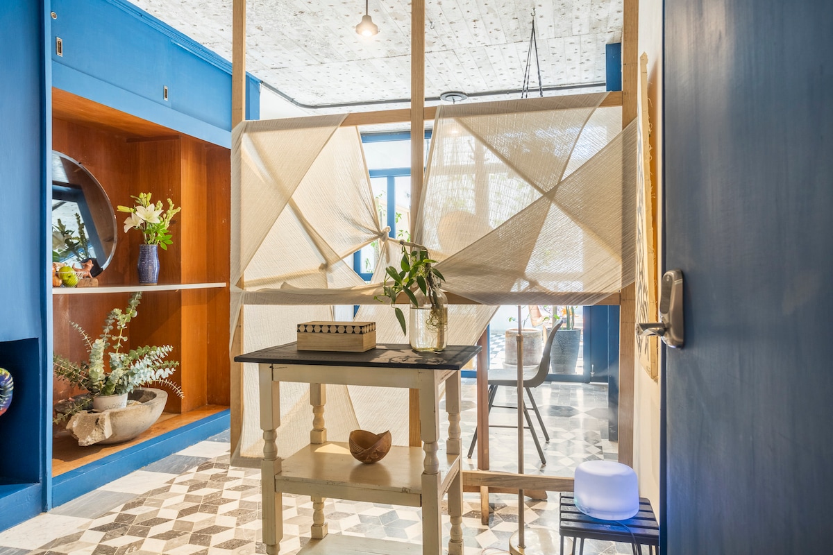 A partition made of natural materials is featured in the foreground, creating a sense of privacy. Behind it, dining and living spaces are visible, showcasing a blend of contemporary design and vibrant colors. Decorative elements and plants enhance the inviting environment.