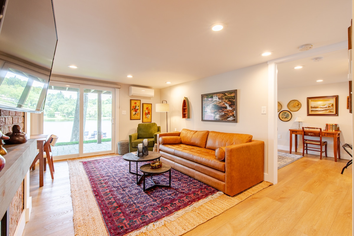 A cozy living area features a brown leather couch and a green armchair, positioned on a light wooden floor. Natural light enters through sliding glass doors, leading to a view of Shickshinny Lake. The room is adorned with artwork and decorative accents, enhancing the space's charm.