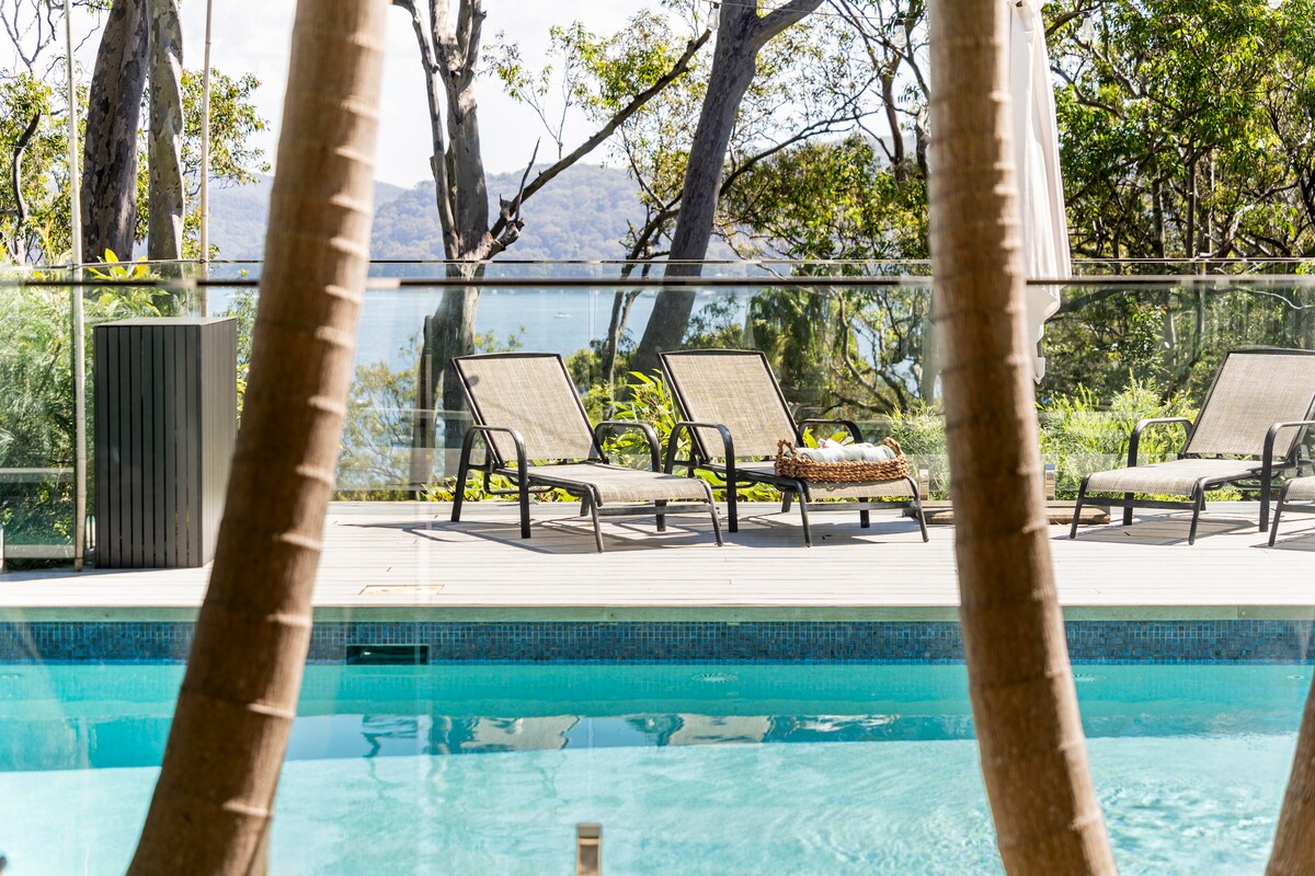 A tranquil outdoor pool is framed by tall trees, offering views of Pittwater in the distance. Sun loungers are positioned alongside the pool, inviting relaxation. The clear blue water reflects the surrounding greenery, creating a serene environment for guests.