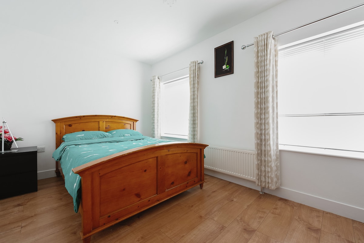 A spacious private room features a wooden bed with a turquoise patterned comforter. Two windows are adorned with light-colored curtains, allowing natural light to enter. A dark wooden nightstand is positioned next to the bed, enhancing the room's simplicity.