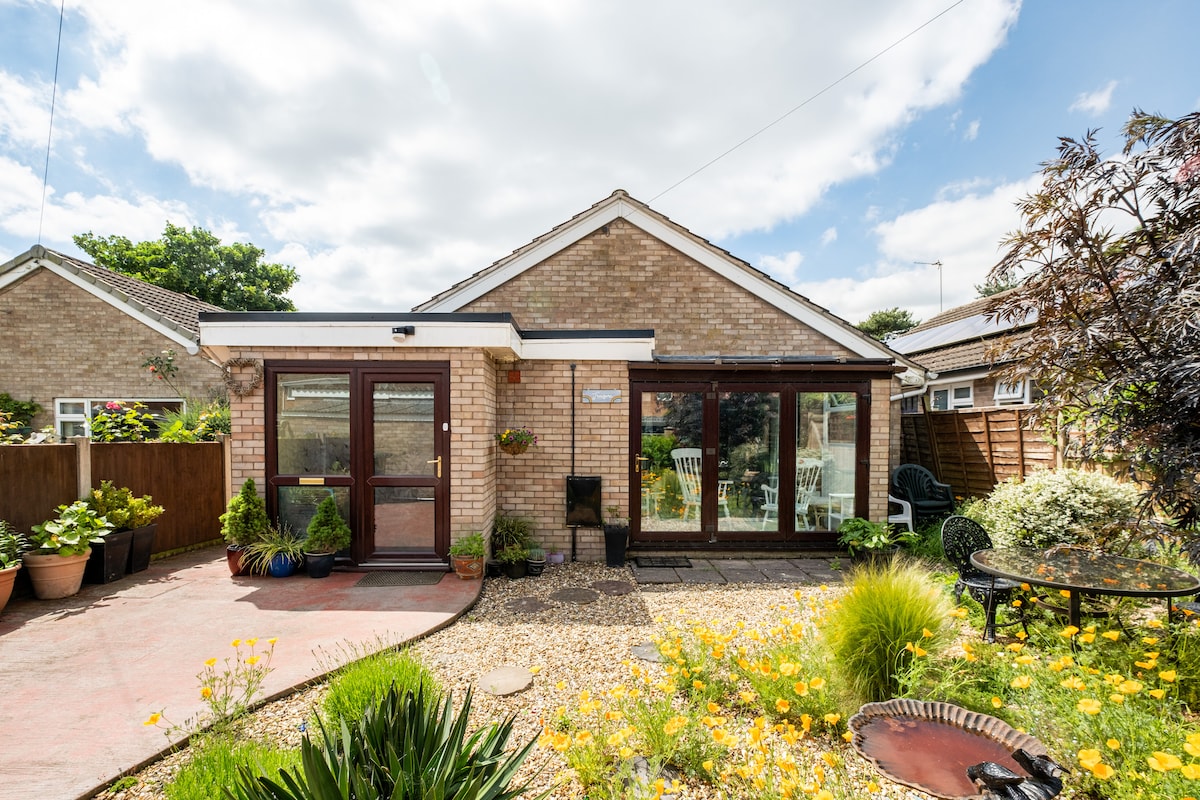 The bungalow's exterior showcases large glass doors leading into a bright entrance, surrounded by a well-maintained garden with colorful flowers and shrubs. A pathway made of stone and gravel guides guests toward the entrance, while outdoor seating is visible in the background.