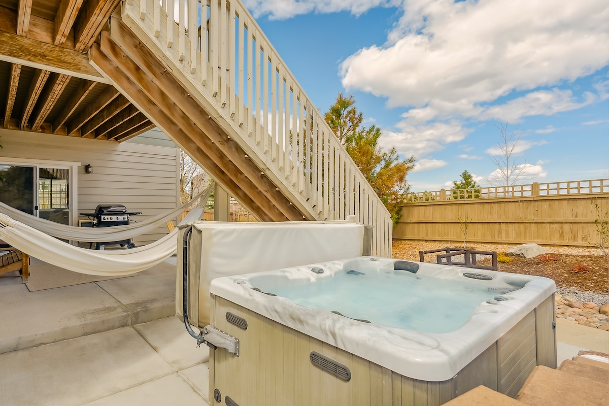 A private hot tub is situated on a patio, inviting relaxation. A barbecue grill is located nearby, alongside a hammock suspended between posts. Steps leading to the upper level are visible in the background, surrounded by landscaped areas and a wooden fence.