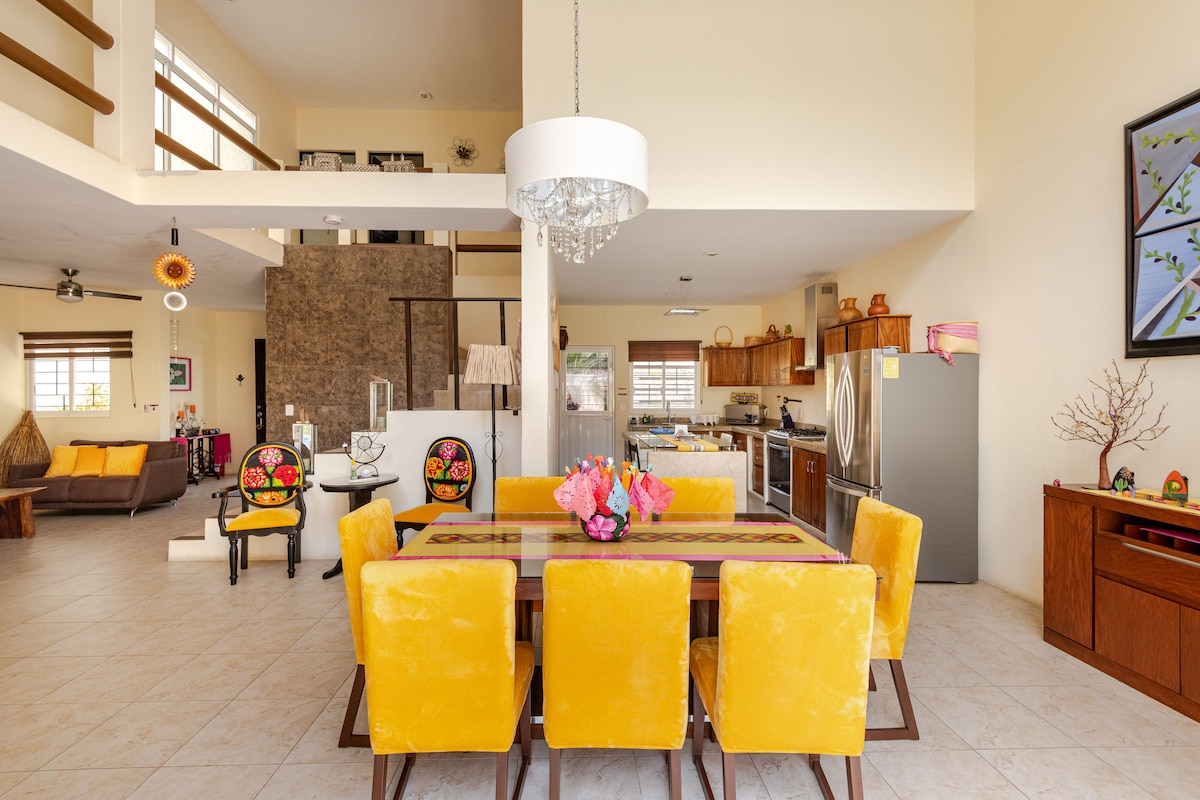 A spacious dining area features a large table surrounded by bright yellow chairs. Natural light floods through tall windows, highlighting the open layout. A decorative chandelier hangs above, while vibrant artwork and decor add color to the walls and surrounding spaces.