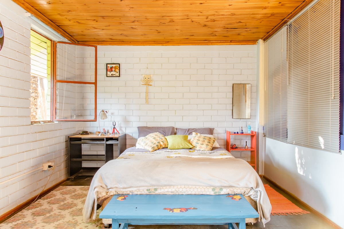 A spacious guest bedroom features a wooden ceiling and light-colored walls. A large bed is positioned against a wall, with pillows placed neatly. A desk and chair are situated nearby, complemented by a small table. Natural light enters through multiple windows, enhancing the room's brightness.