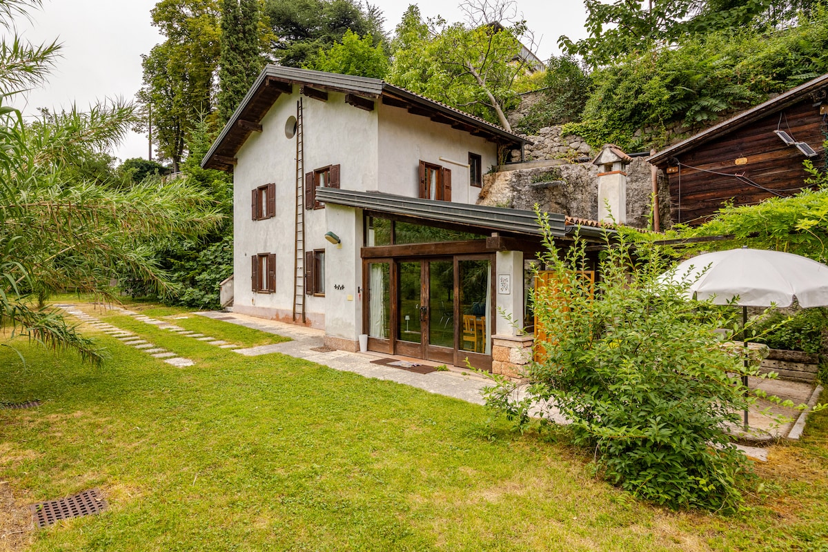 A charming cottage exterior is surrounded by lush greenery, featuring a glass-fronted entrance that invites natural light. A shaded umbrella offers a spot for relaxation, while a stone pathway leads through the garden area. The structure integrates rustic elements with modern design.