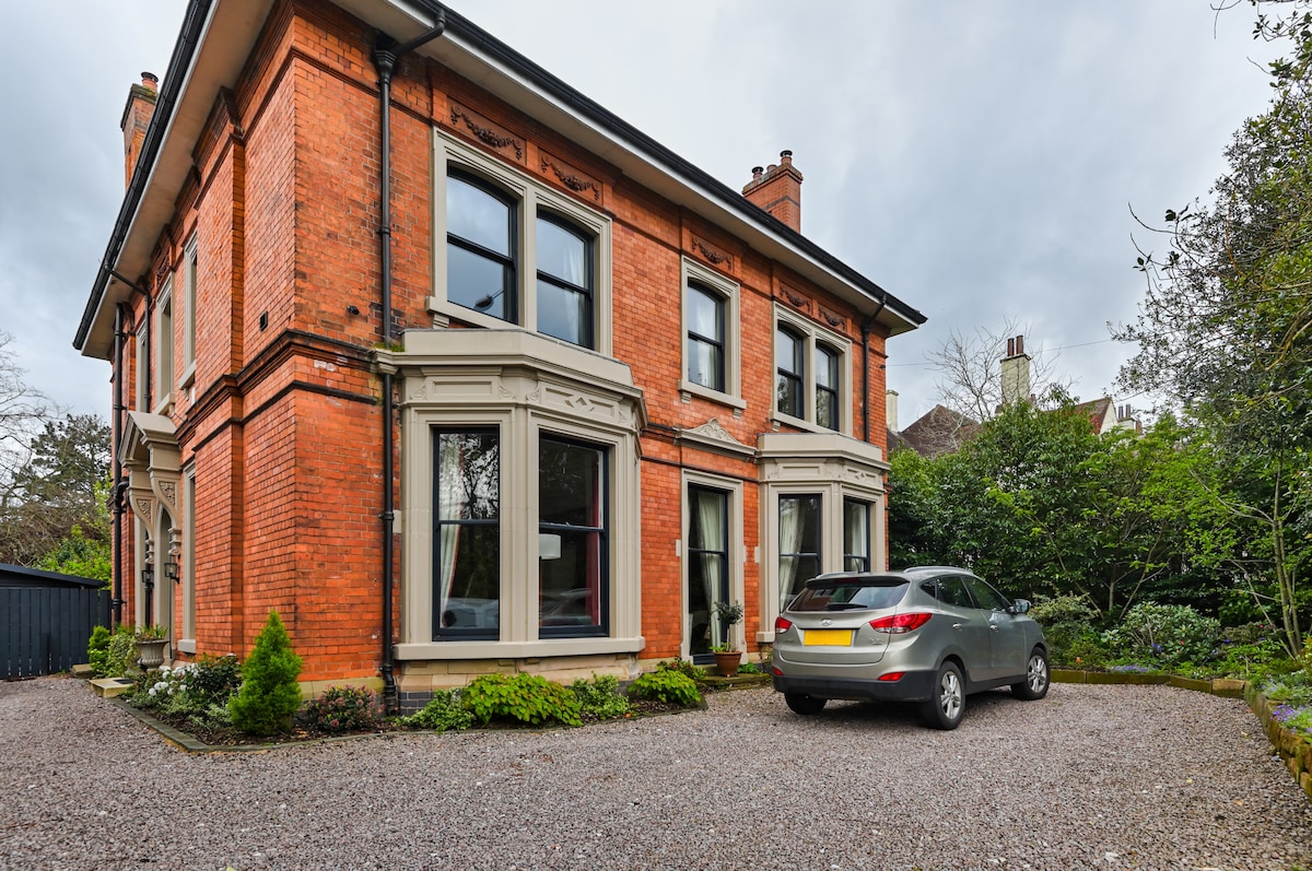 The exterior of a Victorian family home is presented, featuring a red-brick facade and large windows. A gravel driveway is visible, accommodating a parked vehicle. Lush greenery surrounds the property, with shrubs and plants adding to the inviting atmosphere.
