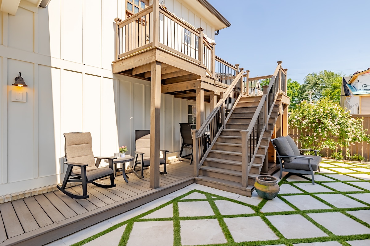 A spacious outdoor area features a wooden deck elevated above the ground, accompanied by a set of stairs. Comfortable seating arrangements and small tables are neatly positioned on the deck. The ground is paved with alternating stone and grassy sections, enhancing the inviting atmosphere.