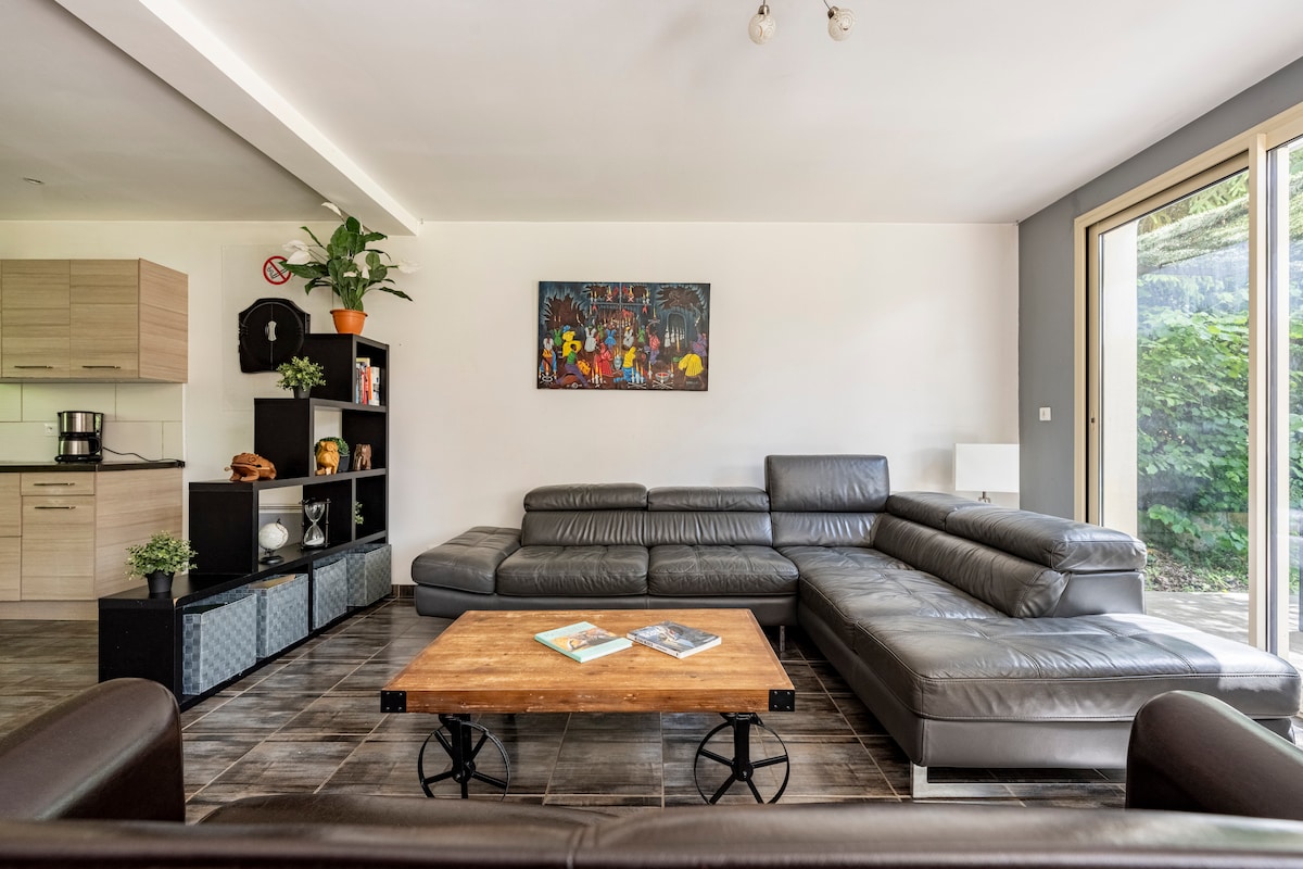 A spacious living area features a large sectional leather sofa and a rustic wooden coffee table. Natural light fills the room through sliding glass doors that open to the outdoor space. A decorative plant and wall art add warmth to the contemporary design.