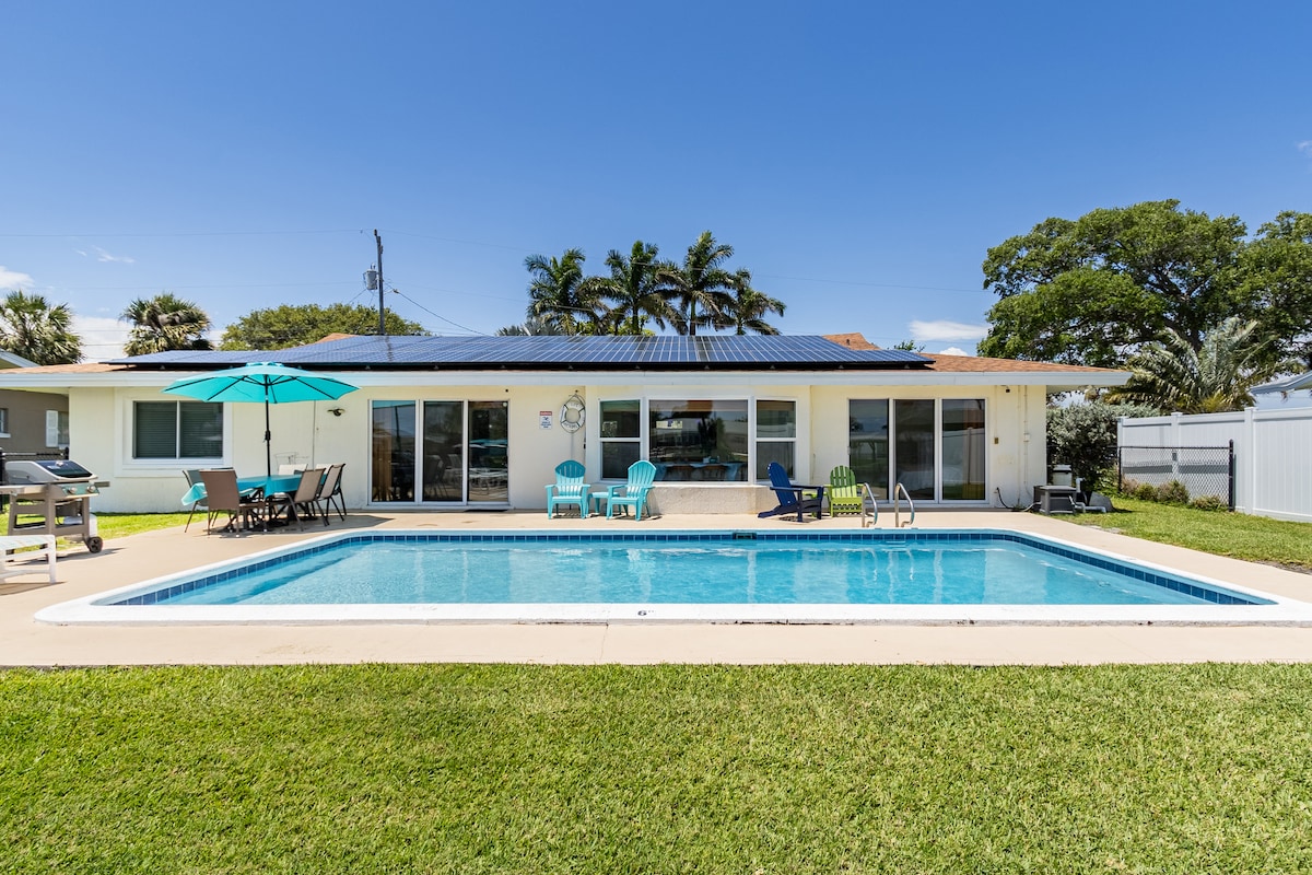 The exterior view showcases a spacious backyard with a sparkling pool surrounded by well-maintained grass. A covered patio with comfortable seating and an umbrella provides shade, while lounge chairs invite relaxation. The house features large glass doors, blending the indoor space with the outdoor area.