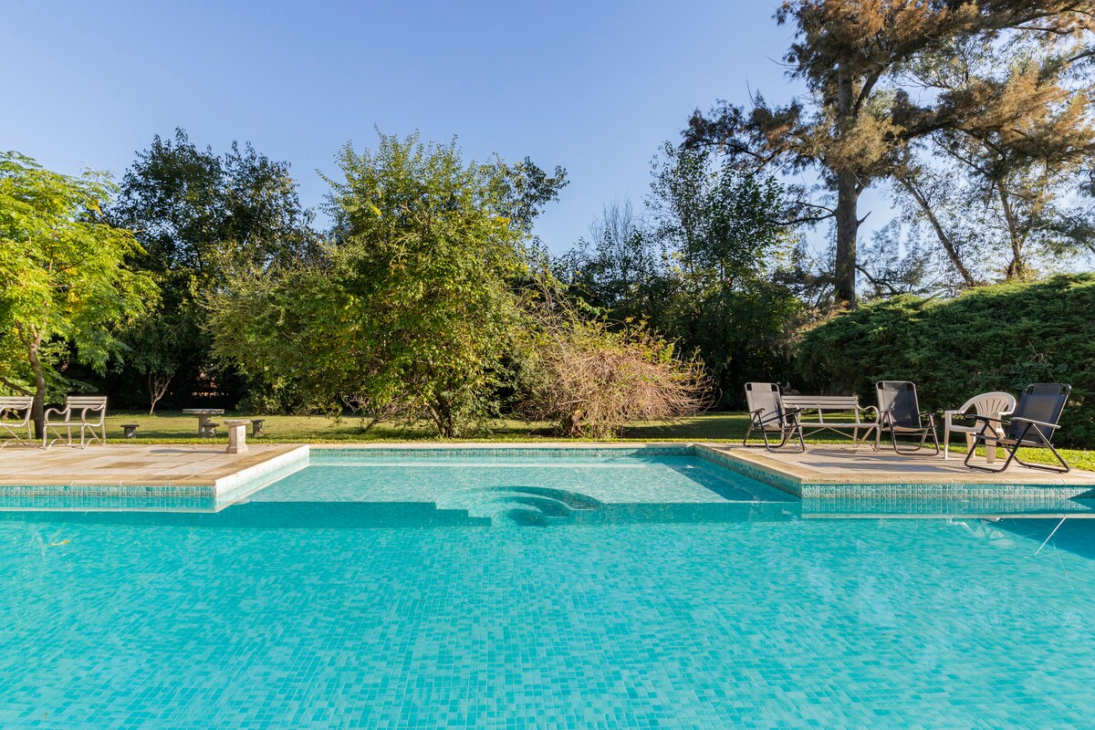 The image shows a clear blue swimming pool surrounded by lush greenery. Lounge chairs are positioned on the wooden deck nearby, offering relaxation space. Trees frames the area, providing a natural backdrop and adding to the serene setting.