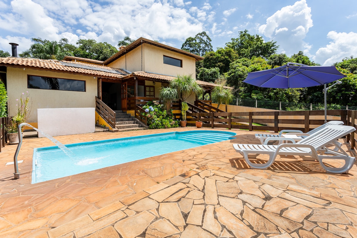 A clear view of the outdoor pool set against a lush green backdrop. The pool area features sun loungers and a shaded umbrella. The textured stone deck surrounds the pool, leading to a well-maintained house with wooden steps and a railing.