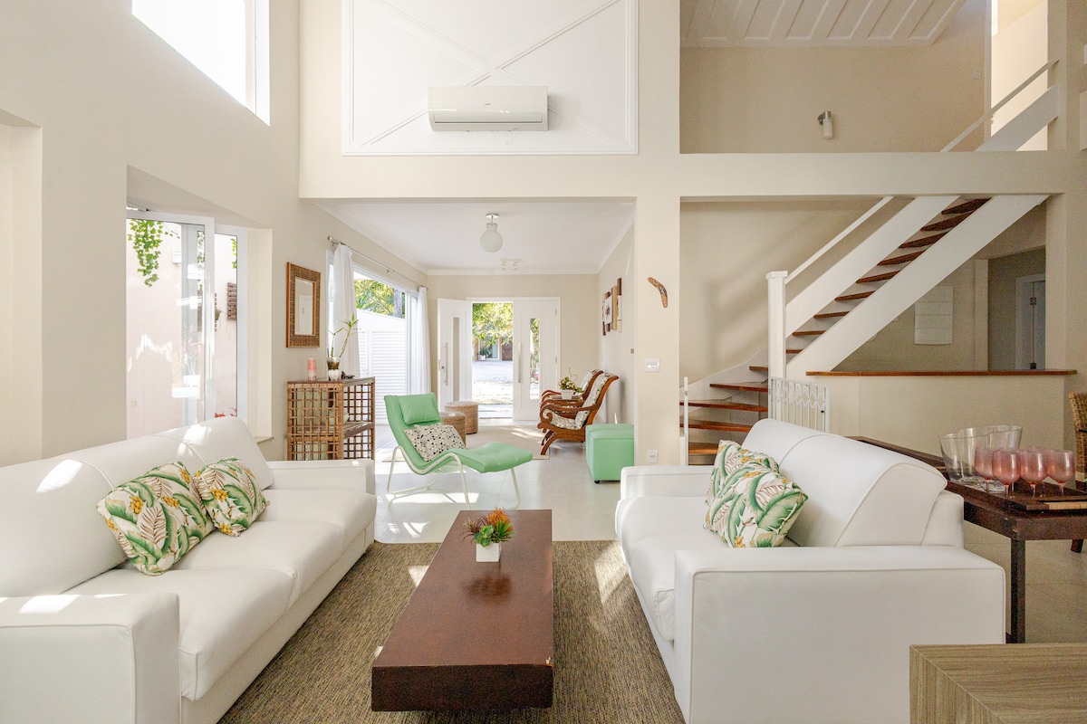 An airy living space features two white sofas adorned with green patterned cushions. A wooden coffee table occupies the center, while large windows allow natural light to fill the room. A set of stairs leads to the upper floor, enhancing the sense of openness.