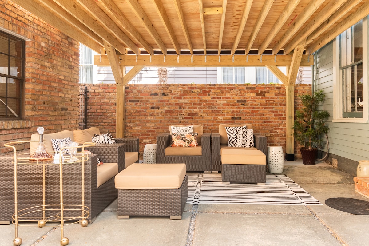 A covered patio space features a relaxing seating arrangement with multiple wicker chairs and a matching ottoman. A soft area rug lies underneath, and decorative cushions add a touch of comfort. Exposed brick walls provide a warm backdrop, enhancing the inviting atmosphere.