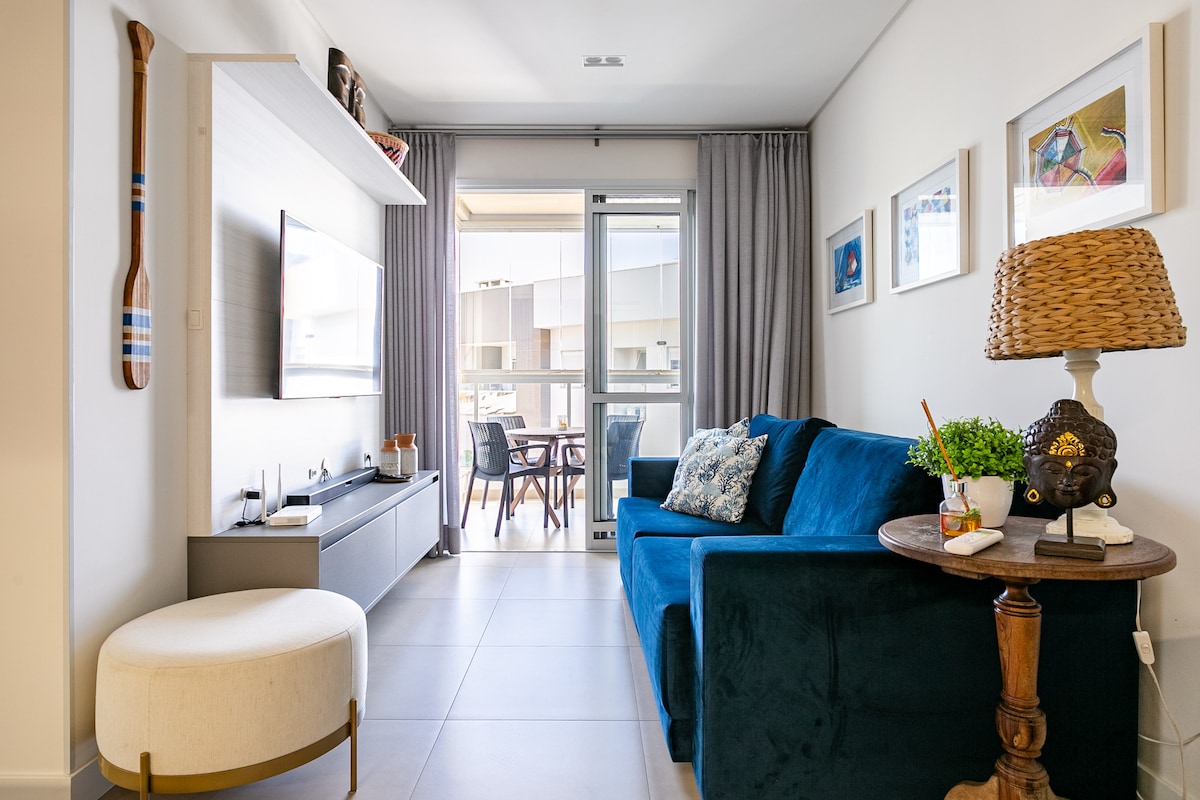 A living area is featured with a plush blue sofa and a decorative table beside it. A television is mounted on the wall, and large glass doors open to a balcony area. Natural light streams in, complemented by soft curtains.