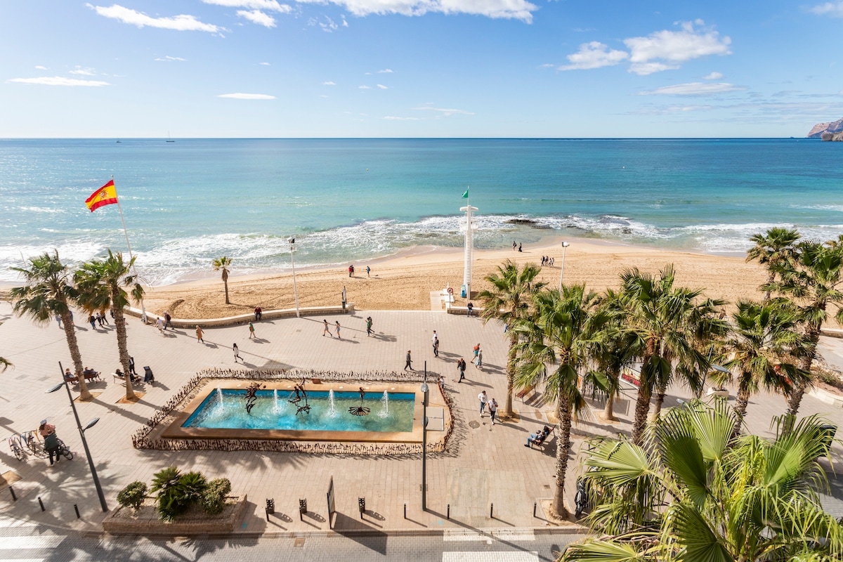 A panoramic view showcases a sandy beach framed by palm trees and a gently waving sea. A fountain sits in the foreground, with a beach promenade bustling with visitors. The sky is bright, dotted with a few clouds, enhancing the coastal atmosphere.