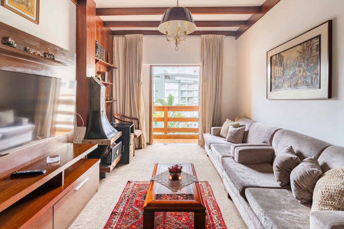 A spacious living area features a comfortable sofa adorned with decorative pillows. A wooden entertainment unit is equipped with a flat-screen TV. Natural light floods through the large window, which is framed by elegant curtains. An intricately designed area rug adds warmth to the room.