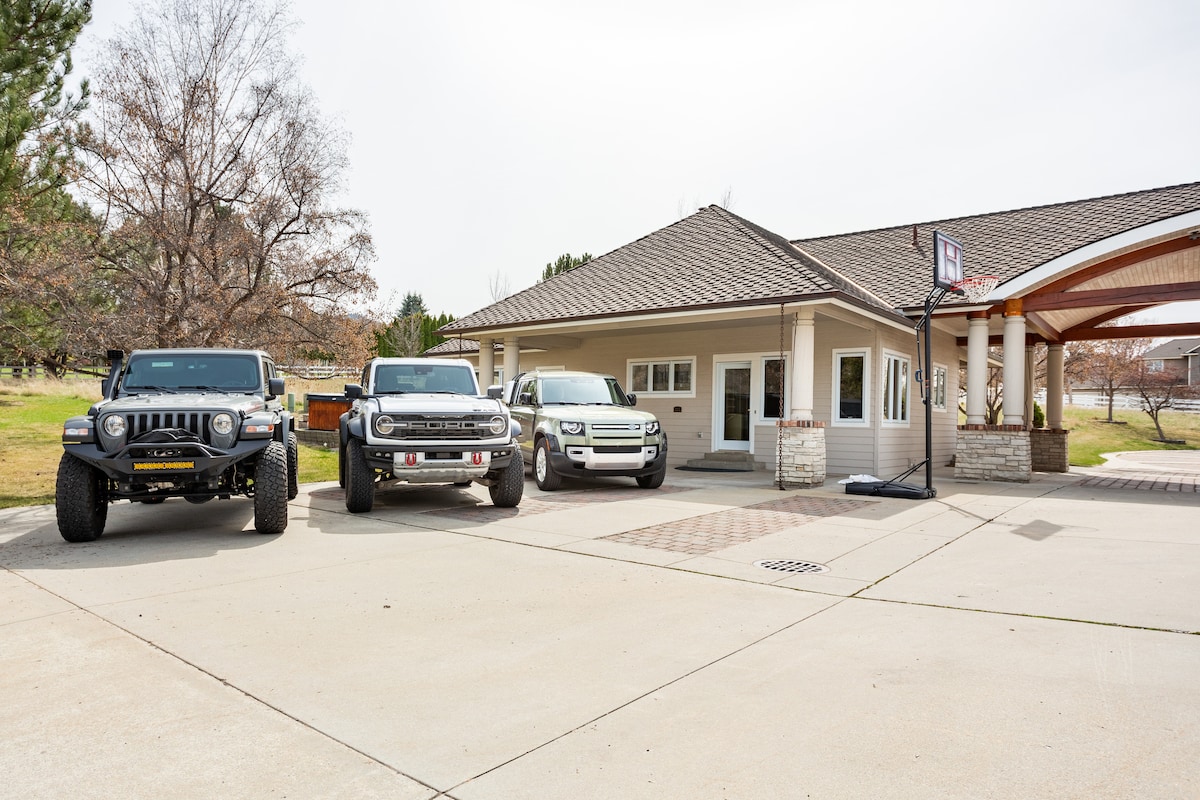 The exterior of a guest home is displayed, featuring a paved driveway with three luxury vehicles parked in front. A welcoming entrance is visible, complemented by landscaped green areas and a basketball hoop nearby. The structure's architecture showcases a covered entry with stone accents.