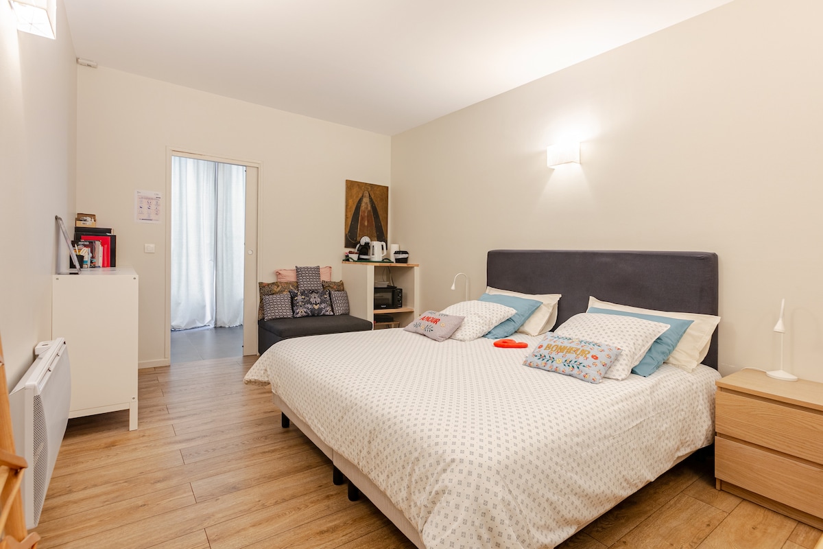 The spacious sleeping area features a comfortable bed dressed in light-colored bedding. A small sofa is positioned nearby, accompanied by a table and a decorative piece on the wall. Soft lighting from wall sconces creates a relaxed ambiance. A doorway leads to another space.