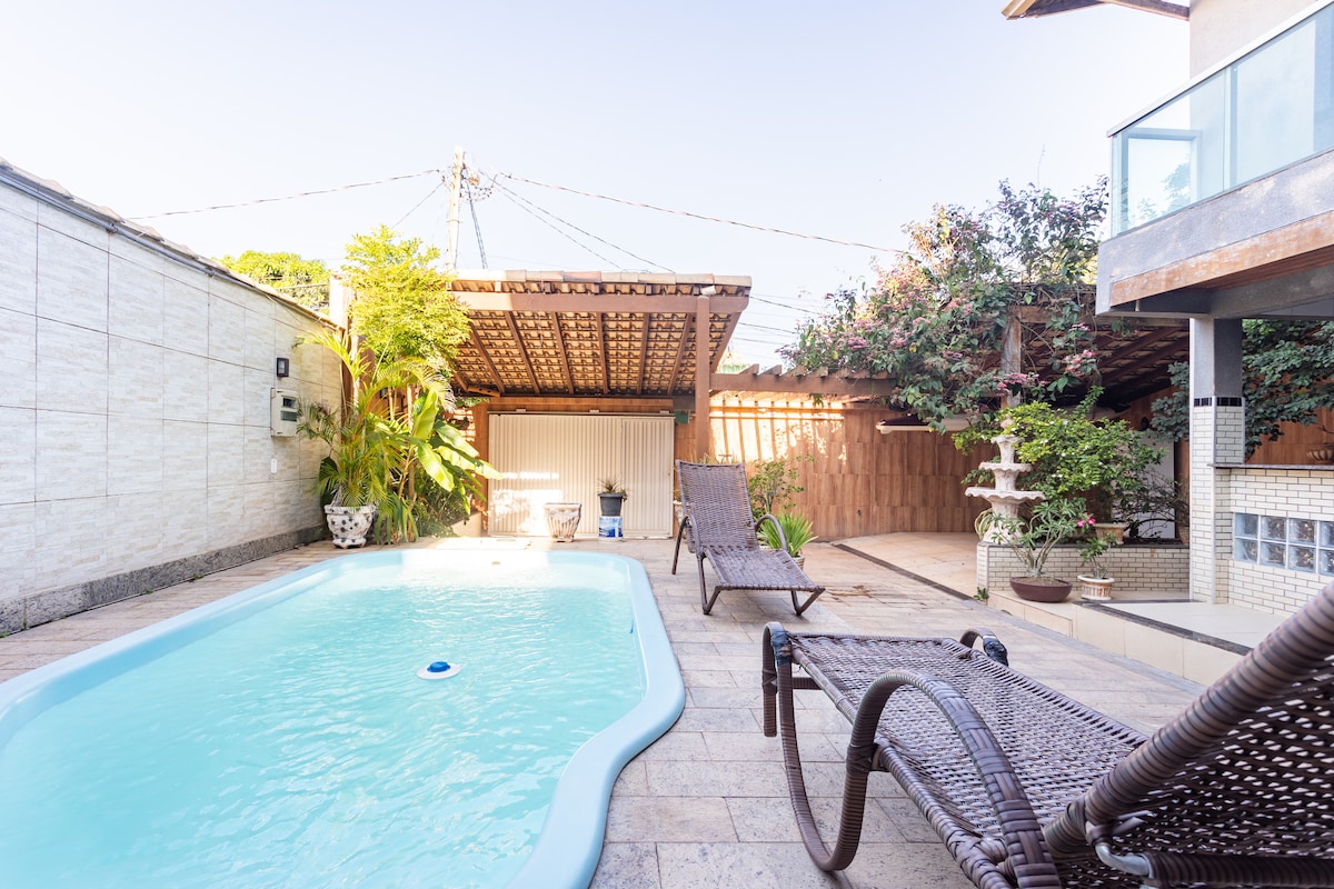 The outdoor area features a refreshing pool surrounded by a spacious patio. Chaise lounges are arranged nearby for relaxation. Lush greenery enhances the ambiance, with structures offering shaded spots, creating a comfortable environment for guests enjoying the serene space.