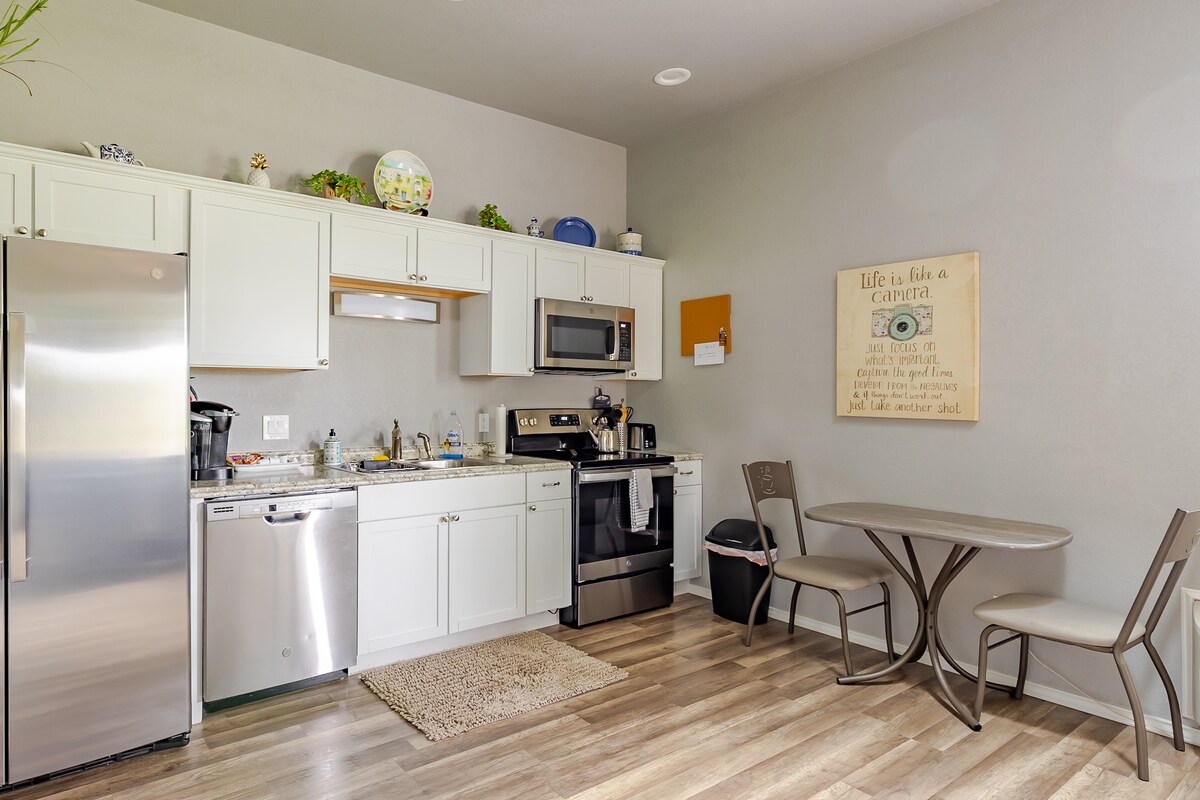 The kitchen area features modern appliances, including a refrigerator, microwave, and oven. A small table is placed nearby with two chairs. Light-colored cabinets provide ample storage. Decorative items are displayed on the shelves, adding a personal touch to the functional space.