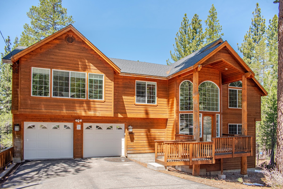 The exterior of a large wooden house is presented, featuring an inviting front porch with steps leading up. Two garage doors are visible, along with numerous large windows offering ample natural light. Surrounding pine trees enhance the peaceful setting.