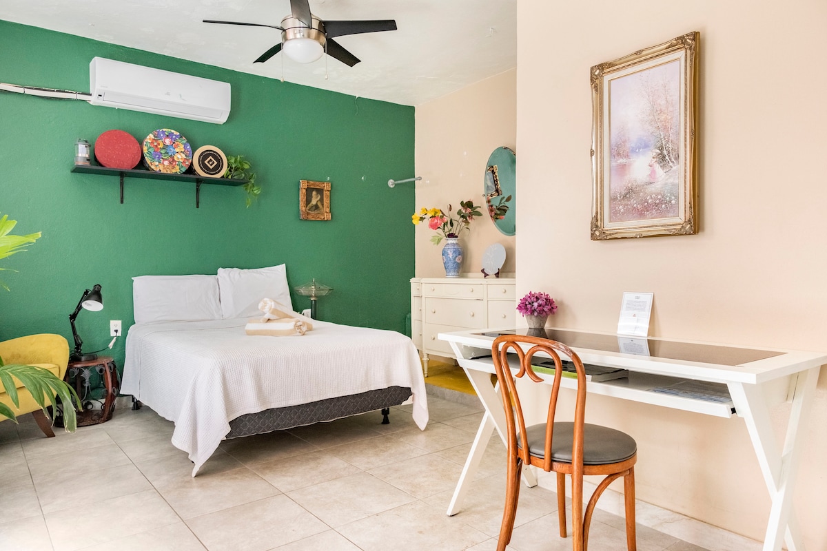 A comfortable sleeping area features a spacious bed with a light-colored blanket. A vibrant green wall provides contrast, while a small desk with a chair is situated nearby. Decorative items adorn the wall shelves, and natural light brightens the space.
