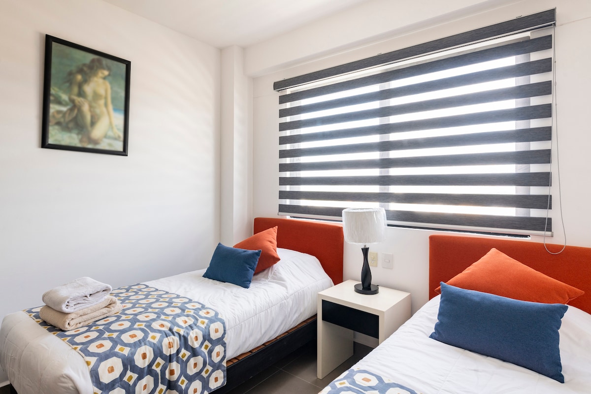 A bright bedroom features two individual beds with orange-styled headboards and contrasting blue cushions. A decorative blanket with a floral pattern rests on one bed. Soft natural light filters through stylish striped window shades, and a small bedside table with a lamp adds functionality.