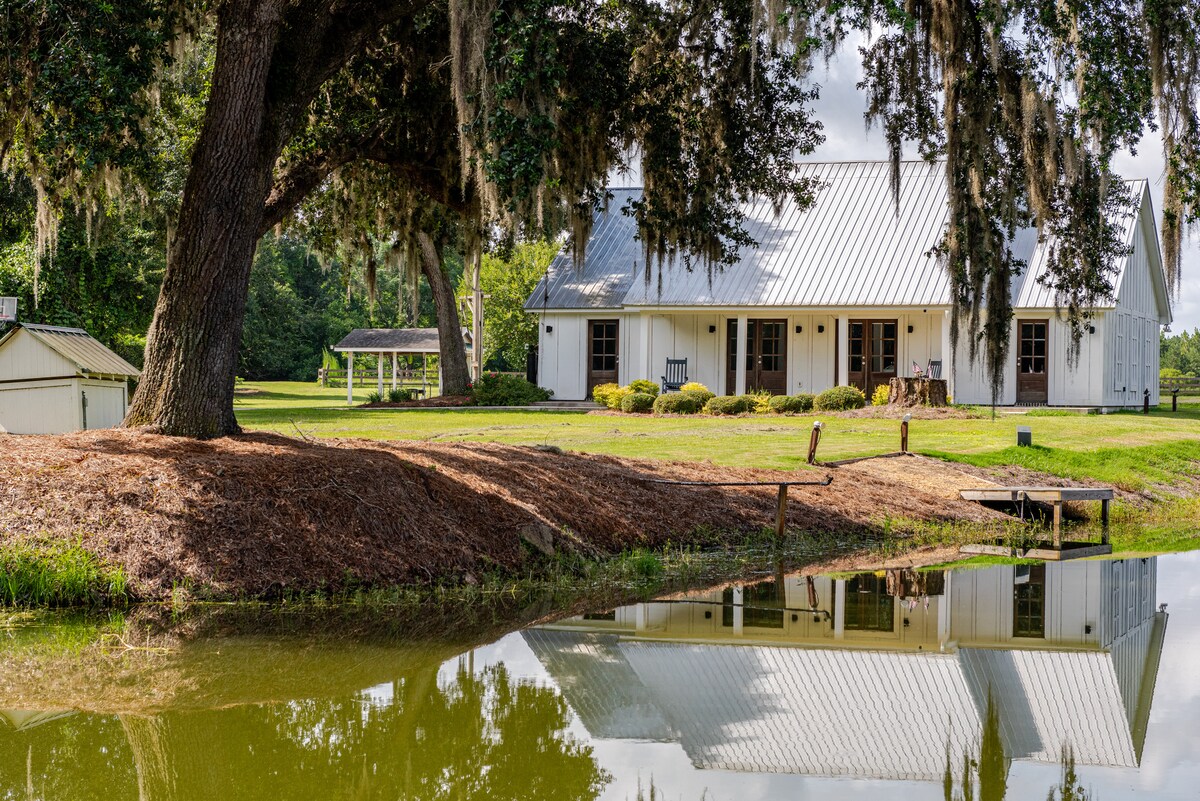 A tranquil farmhouse features a white exterior and a metal roof, set in a green landscape. Reflections can be seen in the calm pond below. Nearby, seating areas and a gazebo are surrounded by lush vegetation, creating a serene outdoor retreat.