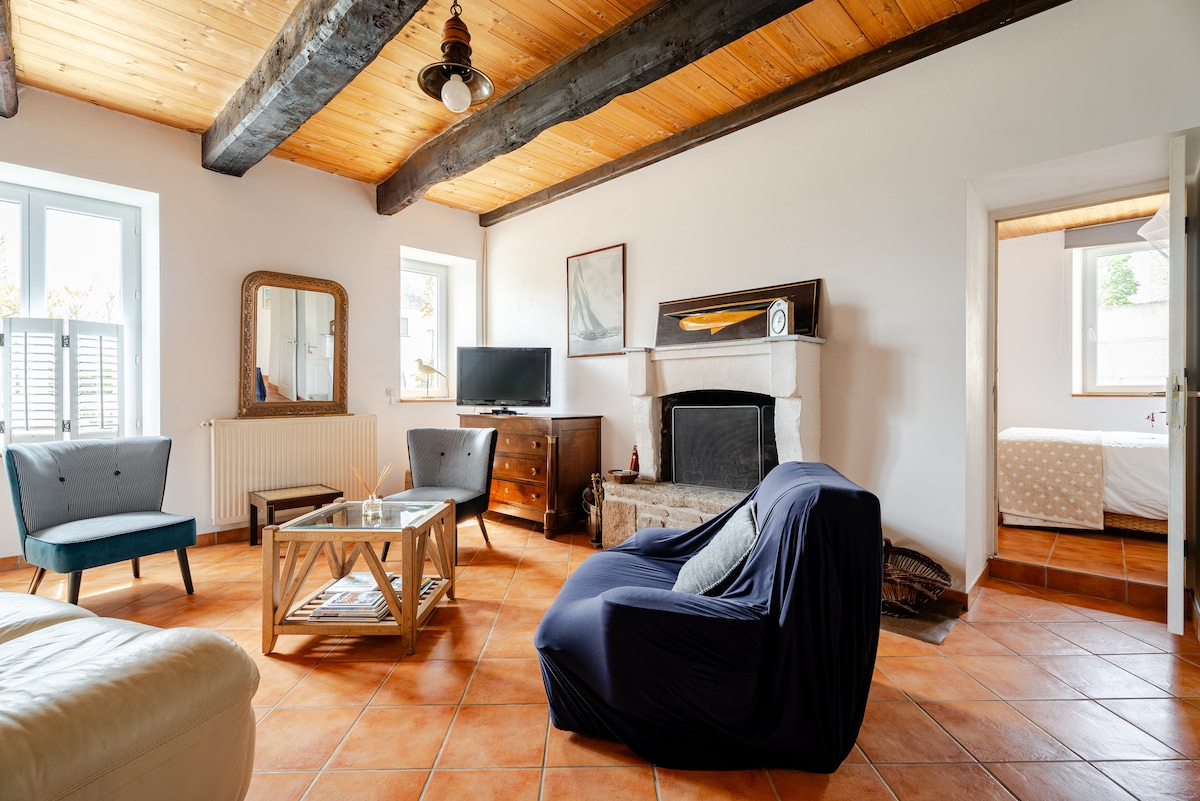 A spacious living area is presented with terracotta tile flooring and exposed wooden beams. A cozy sofa is positioned near a fireplace, accompanied by armchairs and a coffee table. Natural light enters through two windows, enhancing the open feel of the room.