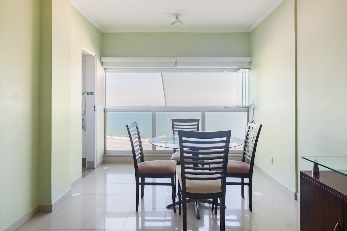 A dining area is furnished with a round table surrounded by four chairs. Large windows offer a view of the ocean, while light green walls create a calm ambiance. A ceiling fan is present, contributing to the airy feel of the space.