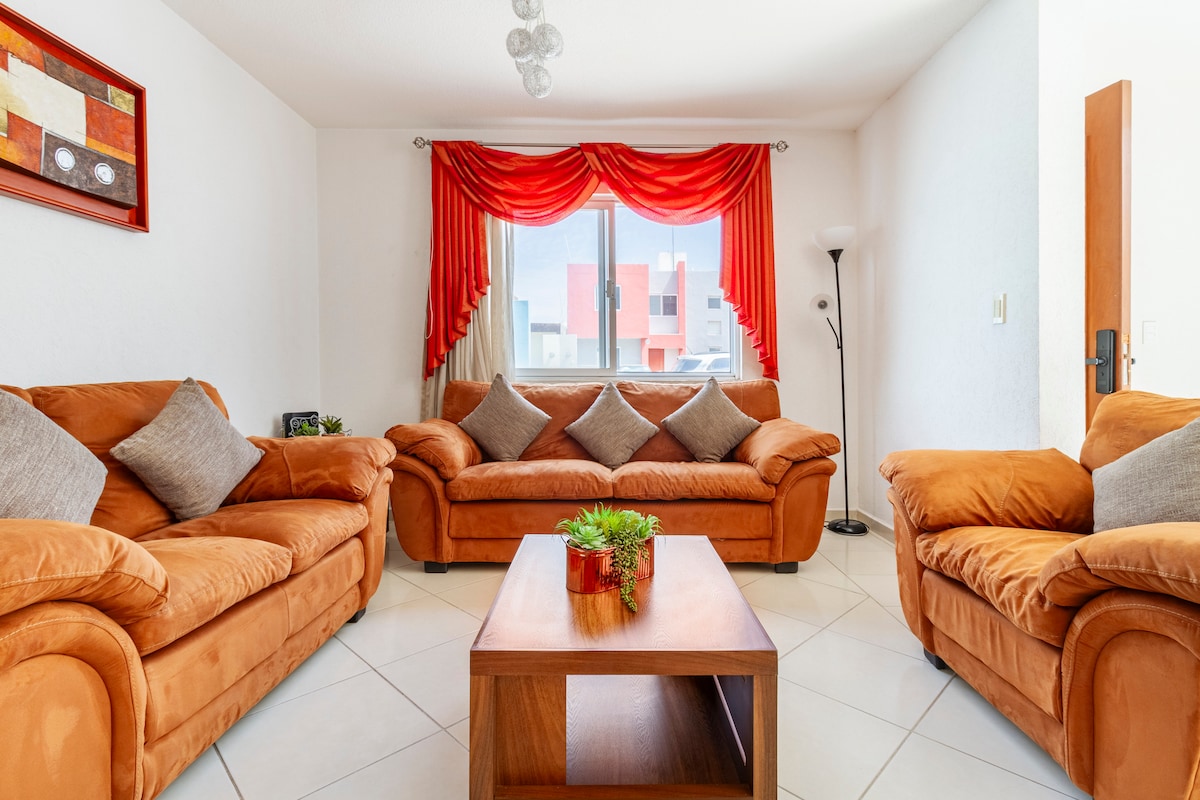 A bright living area features two spacious sofas adorned with decorative cushions, arranged around a wooden coffee table. A large window allows natural light to illuminate the space, complemented by a standing lamp and vibrant curtains.