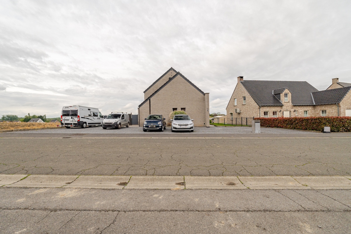 A spacious parking area is visible in front of a two-story building. Several vehicles, including camper vans and cars, are parked along the side of the structure. The surrounding landscape features open fields and a cloudy sky overhead.