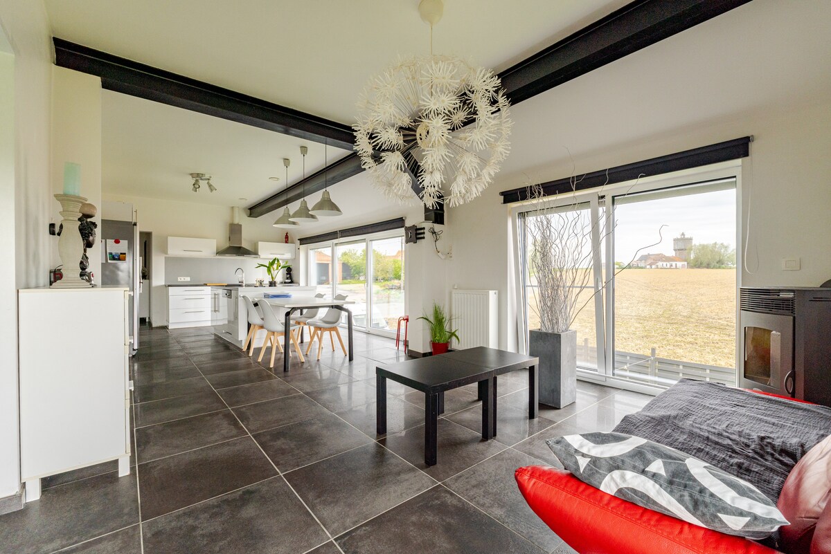 An open living area features a sleek black coffee table at its center, surrounded by a bright red sofa and minimalist decor. Large glass doors invite natural light and offer views of the surrounding fields. The kitchen area is visible, showcasing modern appliances and dining space.