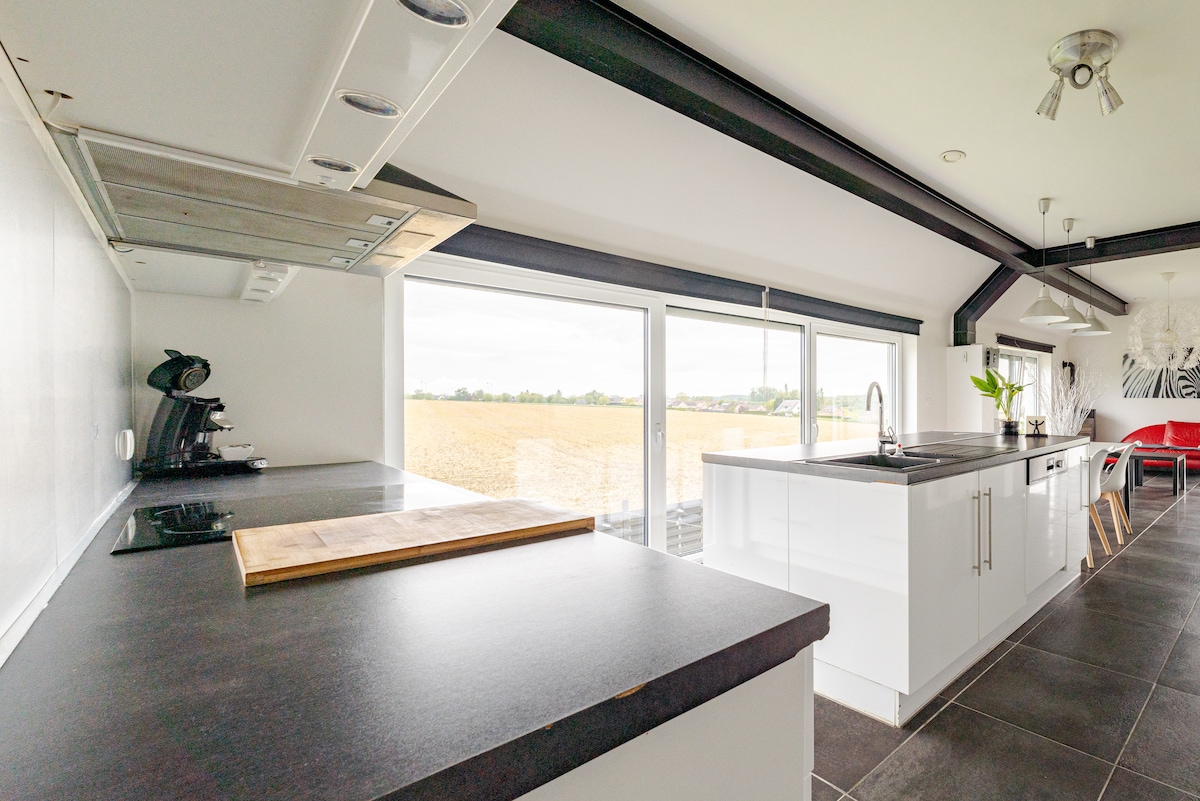 A modern kitchen is featured with sleek black countertops and white cabinetry. Large windows provide ample natural light and an expansive view of the surrounding fields. A coffee maker and stovetop are visible, along with a wooden cutting board on the island.
